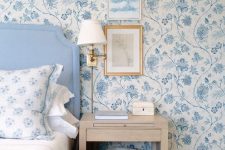 a chic bedroom with blue and white chinoserie-inspired wallpaper, a blue bed with blue and white bedding, a stained nightstand and some artwork