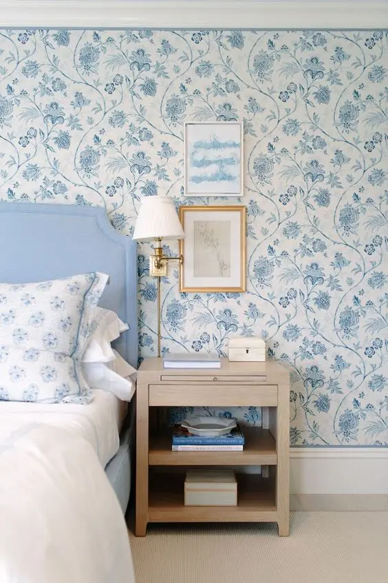 A chic bedroom with blue and white chinoserie inspired wallpaper, a blue bed with blue and white bedding, a stained nightstand and some artwork