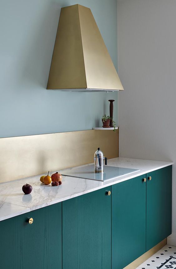 a chic minimal kitchen with emerald lower cabinets, white stone countertops, a gold backsplash and a gold geo hood