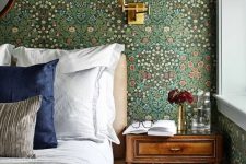 a classic English bedroom with vintage floral wallpaper, an upholstered bed with neutral bedding, a stained nightstand and a sconce