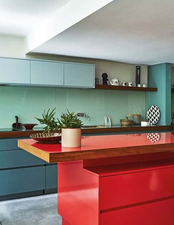 A colorful kitchen with sleek cabinetry in light blue, slate blue and with a bold red kitchen island and a sleek aqua colored backsplash