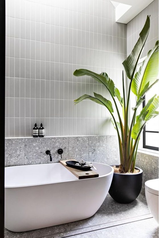 a contemporary bathroom with grey stacked tiles and grey terrazzo ones, an oval tub, a potted plant and some black decor pieces