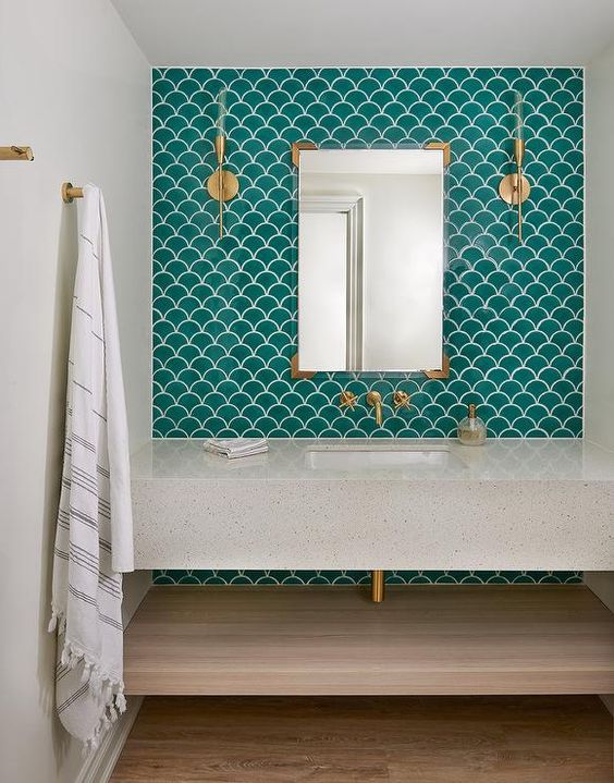 a contemporary neutral bathroom clad with green fishscale tiles, a stone vanity and a shelf and gold fixtures and lights