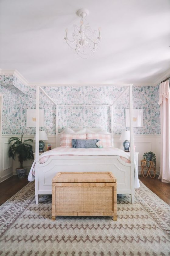 a cottage bedroom with blue floral wallpaper, a white bed with printed bedding, a woven chest, potted plants and a chandelier