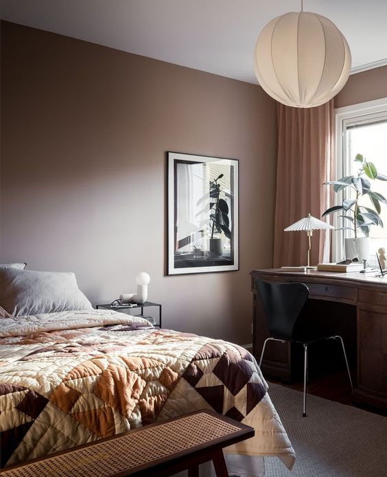 A cozy bedroom with a brown accent wall, a bed with printed bedding, a dark stained desk, a black chair and some plants