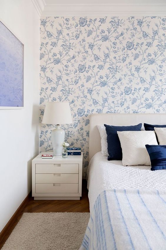 a delicate blue and white bedroom with blue printed wallpaper, a creamy bed and navy and white bedding, creamy nightstands, a rug and an artwork