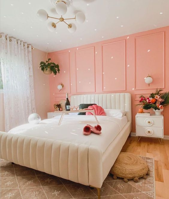 a dreamy bedroom with a peachy pink paneled accent wall, a creamy bed and nightstands, a chandelier and layered rugs