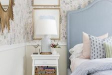a dreamy bedroom with blue floral wallpaper, a blue bed, pastel printed bedding, a white nightstand with a basket and some art