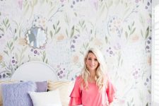 a dreamy pastel bedroom with a bright floral accent wall, a white bed, purple and white bedding, a white nightstand and a pink chandelier