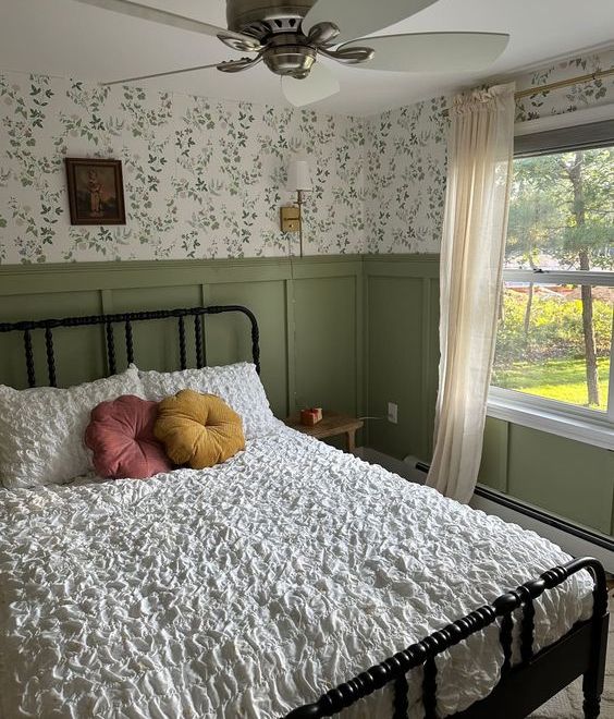 a farmhouse bedroom with green floral wallpaper, green paneling, a metal bed with neutral bedding, bright pillows and some neutral curtains