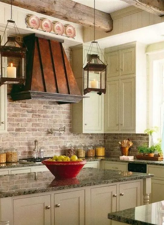 a farmhouse kitchen with shaker cabinets, grey stone countertops, a brick backsplash and a bold aged metal hood that makes a statement