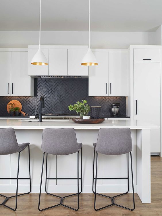 a glam white kitchen with a matte black herringbone tile backsplash, grey stools, white and gold pendant lamps and greenery
