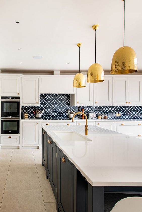 a glam white kitchen with shaker cabinets, a navy kitchen island, white stone countertops, a navy fish scale tile backsplash and gold pendant lamps