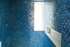 a gorgeous bathroom clad with blue fishcale tiles all over, with a white floor and a toilet, with a window and a niche shelf