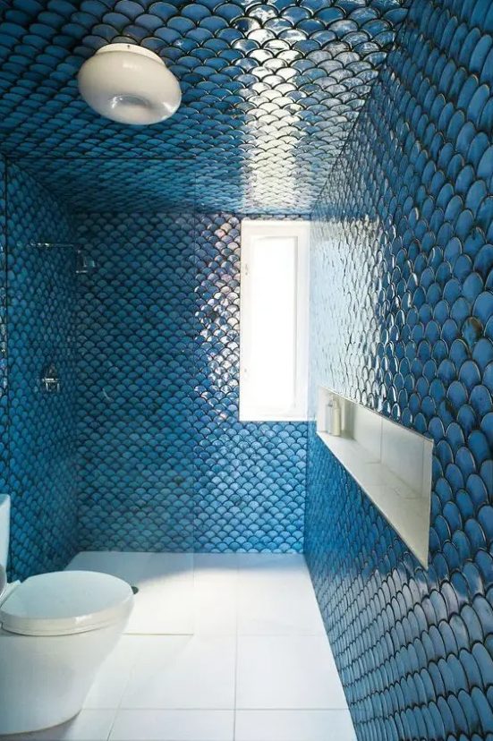 a gorgeous bathroom clad with blue fishcale tiles all over, with a white floor and a toilet, with a window and a niche shelf