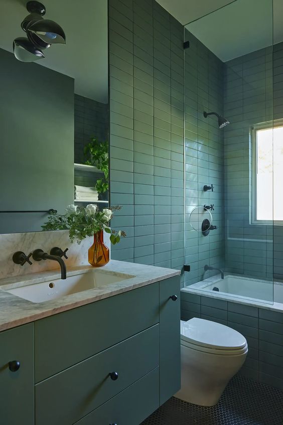 a green bathroom clad with stacked tiles, a tub with a divider, a green vanity and a large mirror plus black fixtures
