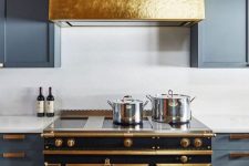 a grey kitchen with shaker cabinets, white stone countertops and a backsplash, a shiny gold hood over a chic vintage cooker
