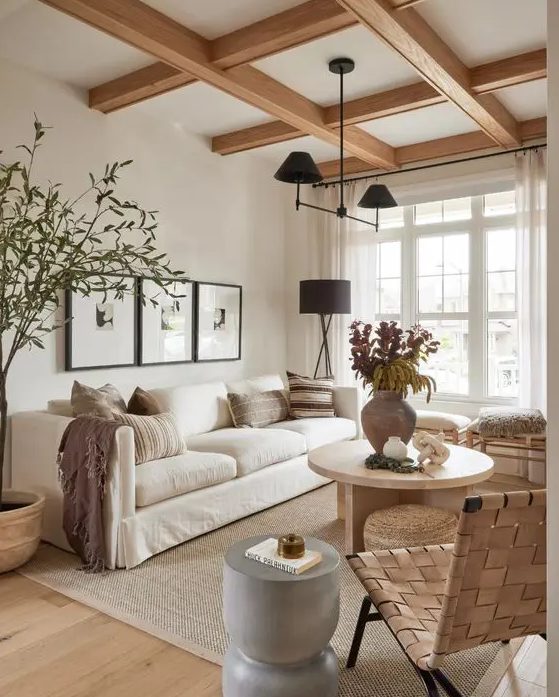 A light filled earthy living room with wooden beams, a white sofa, stools and a woven chair, a grey side table and a round coffee table