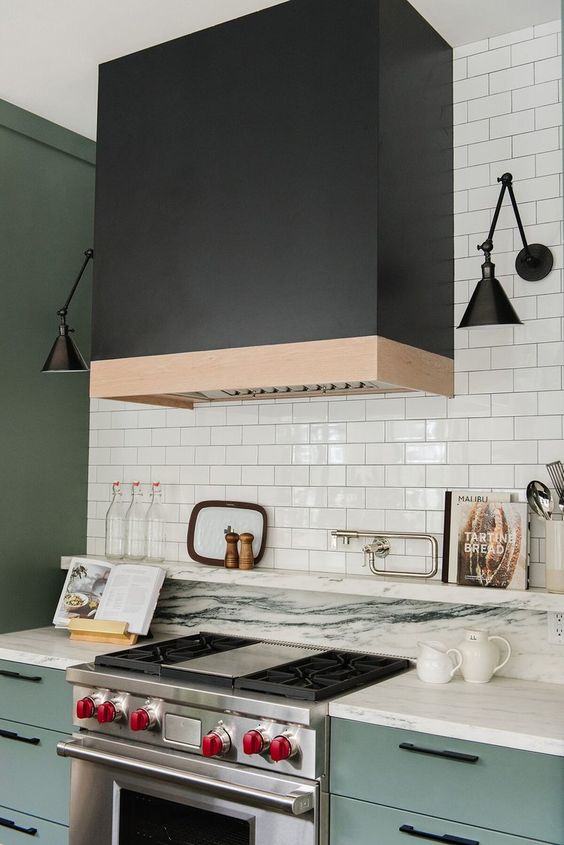 a light mint green kitchen with a white tile backsplash and countertops and an oversized black hood plus black sconces