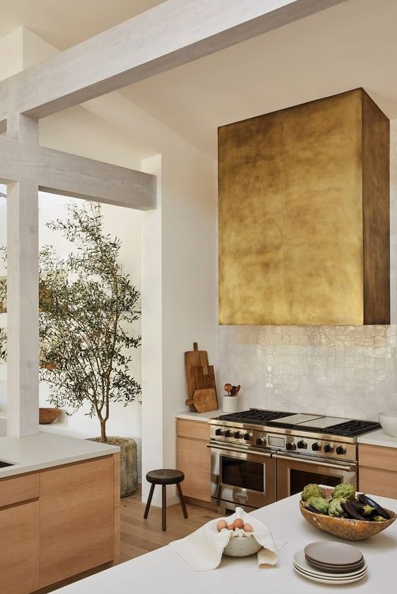 A light stained kitchen with a white Zellige tile backsplash and a gilded oversized hood is a catchy and cool space