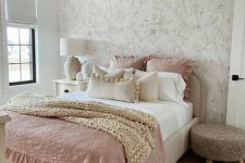 a lovely and delicate bedroom with a floral accent wall, a neutral bed with pastel and neutral bedding, a white nightstand, a woven pouf