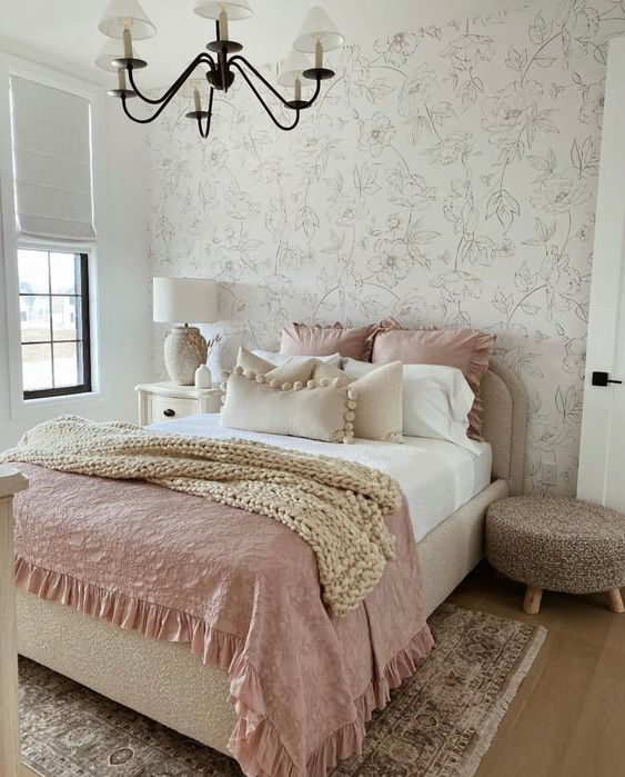 a lovely and delicate bedroom with a floral accent wall, a neutral bed with pastel and neutral bedding, a white nightstand, a woven pouf