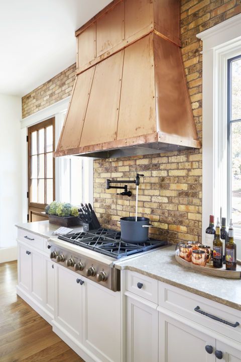 a lovely white farmhouse kitchen with shaker cabinets, a brick backsplash and a large copper hood that adds a warm touch