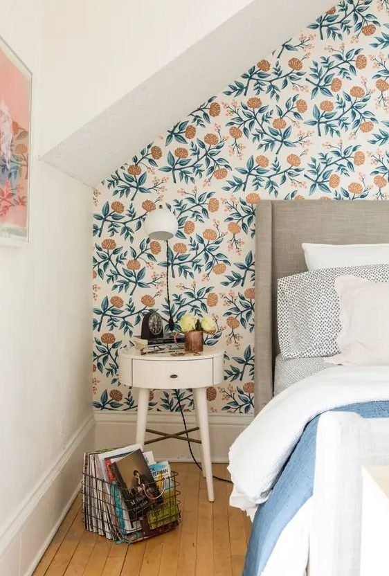 A mid century modern bedroom with floral wallpaper, a greige bed with neutral and blue bedding, a nightstand with a lamp