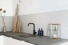 a minimalist Scandinavian kitchen with sleek white cabinets, concrete countertops, a grey herringbone tile backsplash, pendant bulbs