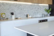 a minimalist kitchen with stained and white cabinets, white countertops, a marble scallop tile backsplash and lights