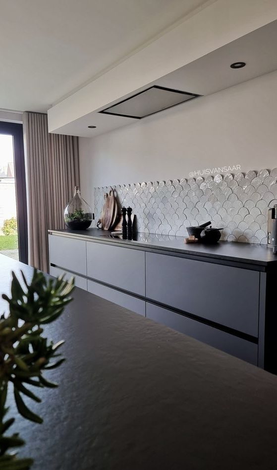 A minimalist sleek grey kitchen with only lower cabinets, a white fishscale tile backsplash and a built in hood over the space