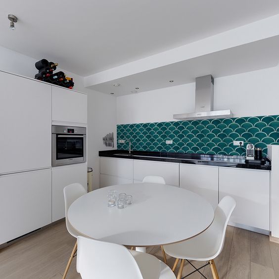 a minimalist white kitchen with sleek cabinets, black countertops, an emerald fish scale tile backsplash and a dining set