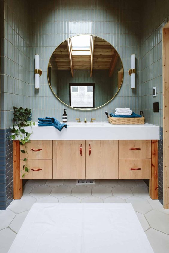 A modern bathroom clad with green stacked and light hex tiles, a light stained vanit and a round mirror