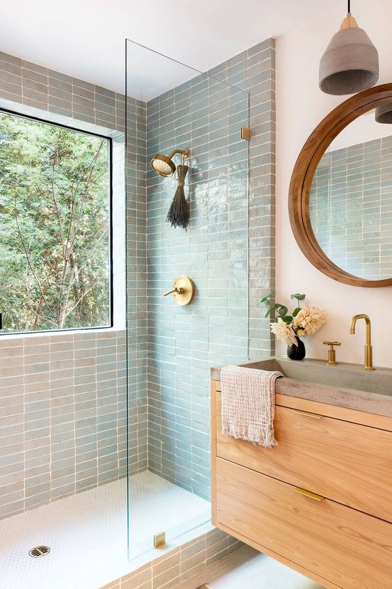 a modern bathroom with blue green stacked tiles in the shower, a light-stained vanity, a mirror with a wooden frame