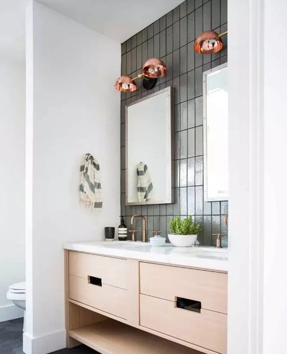 a modern bathroom with grey stacked skinny tiles, a blonde wood vanity with a white countertop, brass fixtures and copper sconces