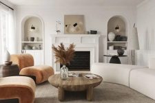 a modern earthy living room with a fireplace, rust-colored chairs, a neutral curved sofa, a low coffee table and niches with decor
