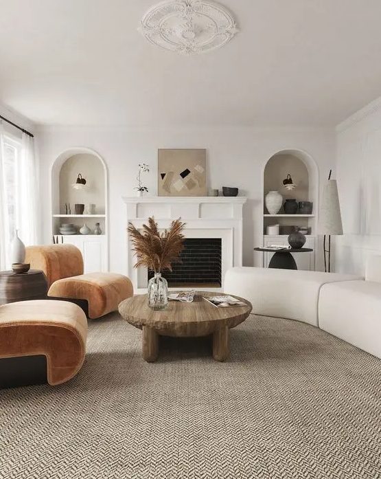 A modern earthy living room with a fireplace, rust colored chairs, a neutral curved sofa, a low coffee table and niches with decor