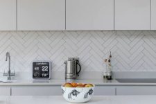 a modern kitchen with sleek white cabinets and a grey kitchen island, a white herringbone tile backsplash and pendant bulbs