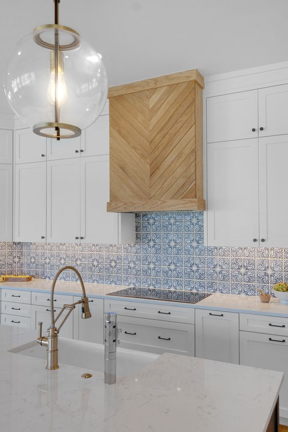 a modern white kitchen with shaker cabinets, a blue printed tile backsplash, a light-stained wooden hood for a warm touch