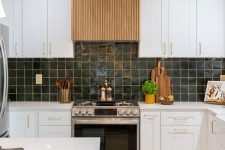 a modern white kitchen with shaker cabinets, a fluted wood hood, a dark green Zellige tile backsplash and lights