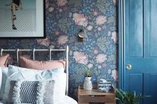 a moody bedroom with dark floral wallpaper, a metal bed with pastel bedding, a stained nightstand, some art and plants