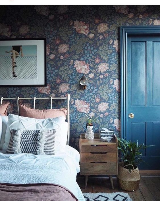 a moody bedroom with dark floral wallpaper, a metal bed with pastel bedding, a stained nightstand, some art and plants