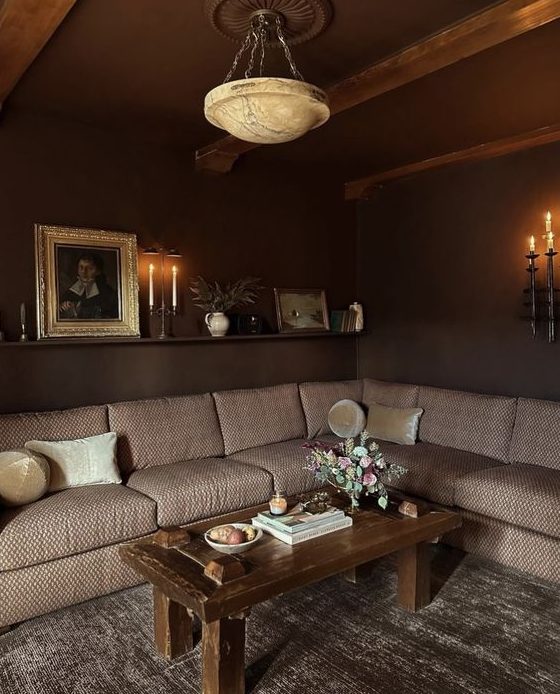 a moody living room with dark brown walls, a beige sectional, a coffee table and lovely art adn blooms