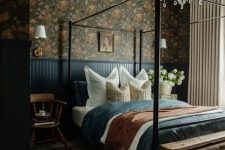 a moody vintage bedroom with dark floral wallpaper, a metal canopy bed, navy paneling and bedding, a crystal chandelier
