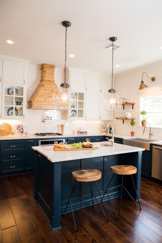 A navy and white kitchen with a white tile backsplash and countertops, a light stained wooden hood for an accent