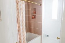 a neutral bathroom with pink stacked tiles in the bathtub space, white herringbone ones on the floor and gold touches