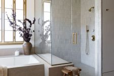 a neutral earthy bathroom with a herringbone floor, neutral tiles, a bathtub in a stone slab and some wooden touches
