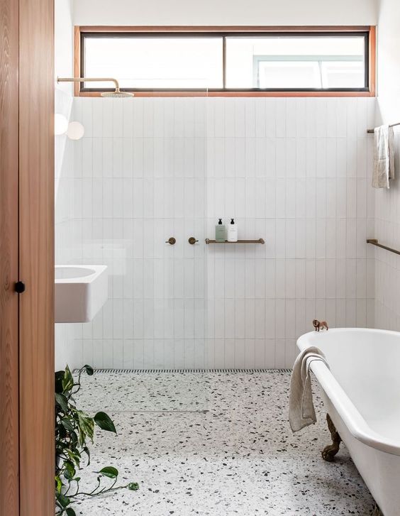 a neutral modern bathroom clad with white stacked tiles, white terrazzo, a free-standing tub and greenery