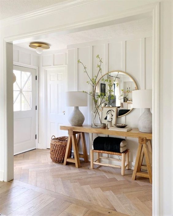 A neutral modern farmhouse space with a light stained herringbone floor, a trestle console table, a stool and a large mirror, some lamps