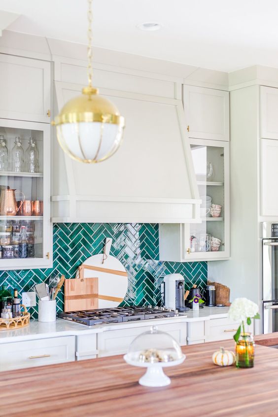 a pale green kitchen with shaker and glass cabinets, a large hood, an emerald herringbone tile backsplash, a gold pendant lamp